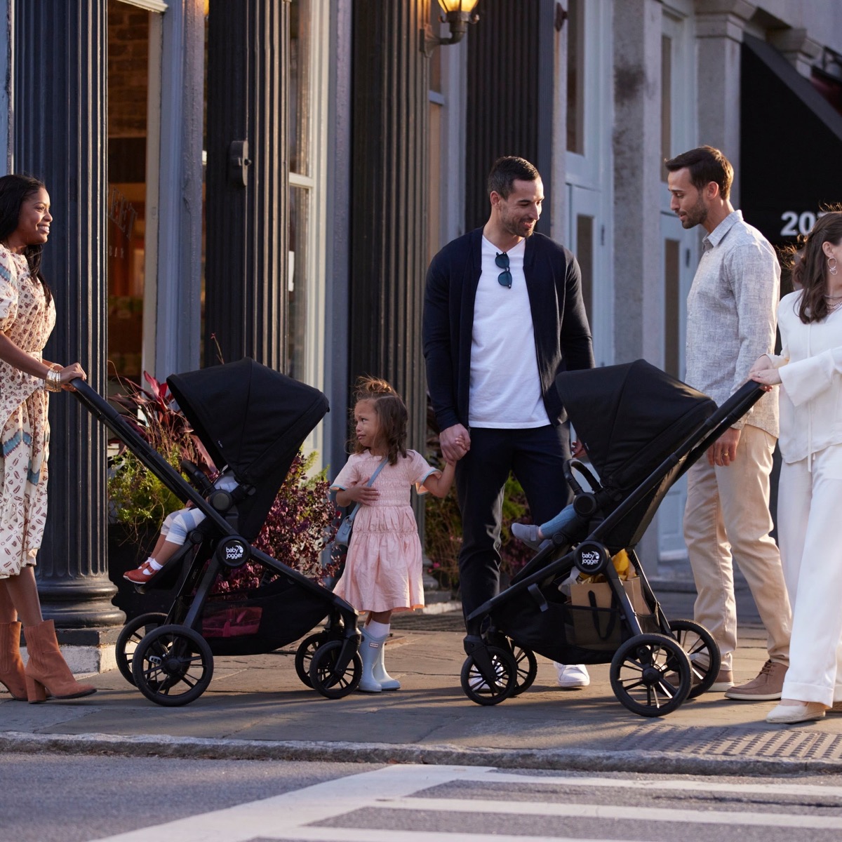 Group lifestyle photo on a city sidewalk showing two Baby Jogger strollers being pushed, one with a child riding