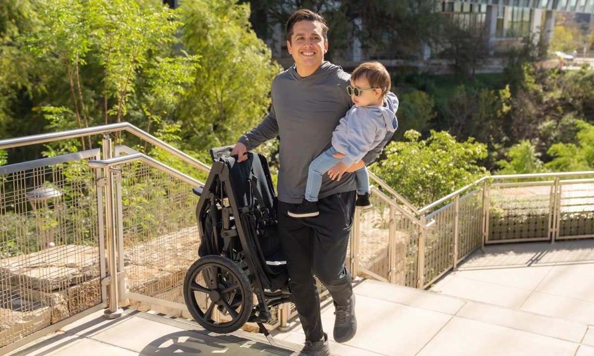 Caregiver holding a child next to a folded Wayfinder jogging stroller in the Nightfall fashion, showing the space-saving DualRetract fold