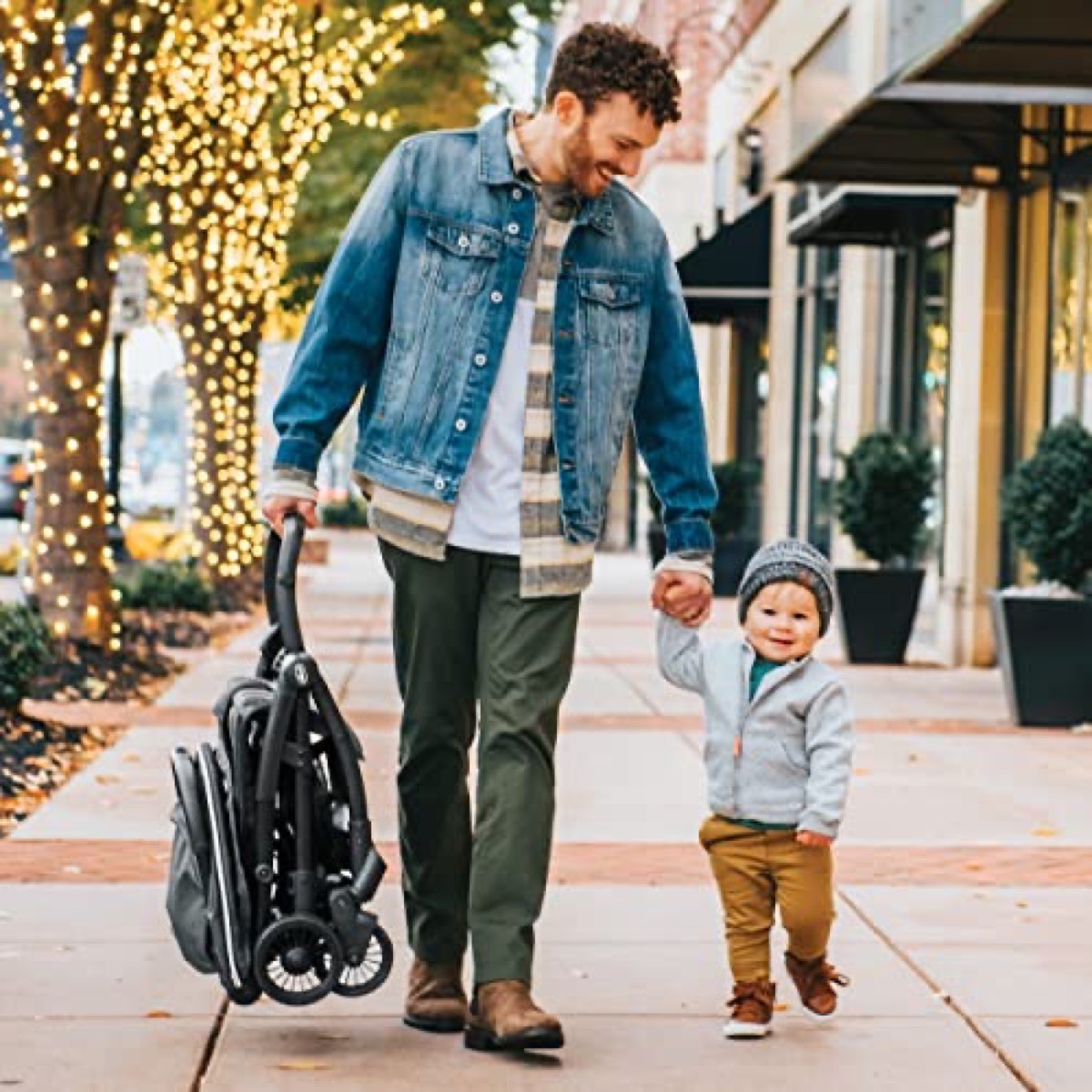 Adult walking on a city sidewalk holding a toddler’s hand while rolling the folded Chicco Presto beside them, showing its compact folded footprint