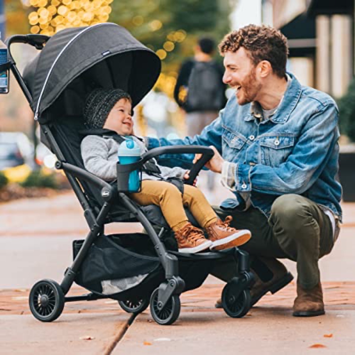 Toddler seated in the unfolded Chicco Presto with a bottle in the armbar cup holder while an adult crouches beside the stroller on a sidewalk