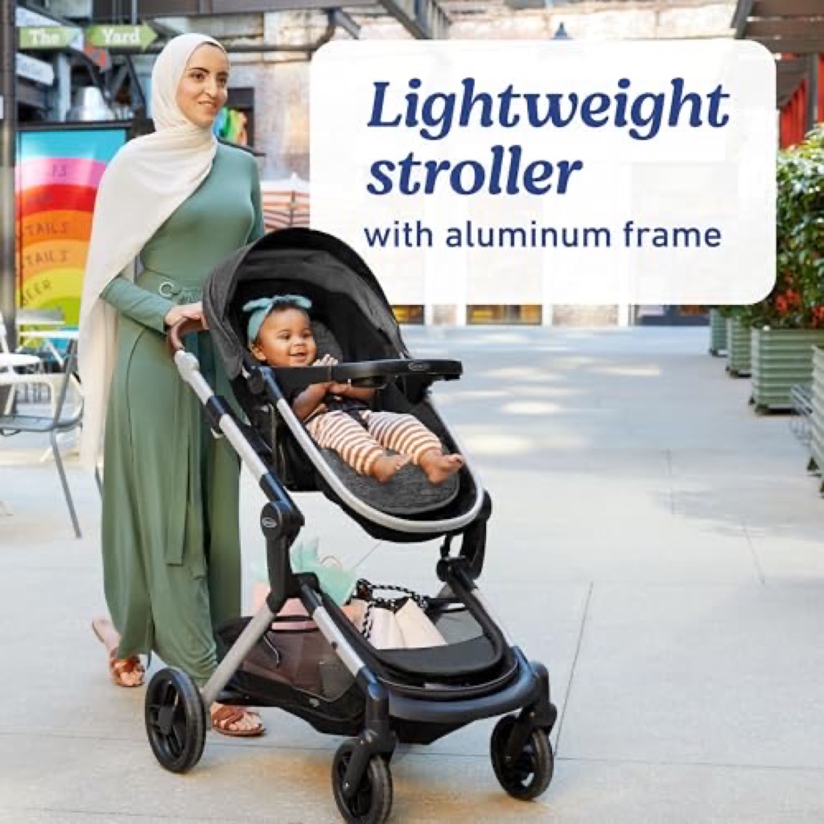 A caregiver in a green abaya pushing a toddler in the charcoal Modes Nest stroller along a paved city sidewalk