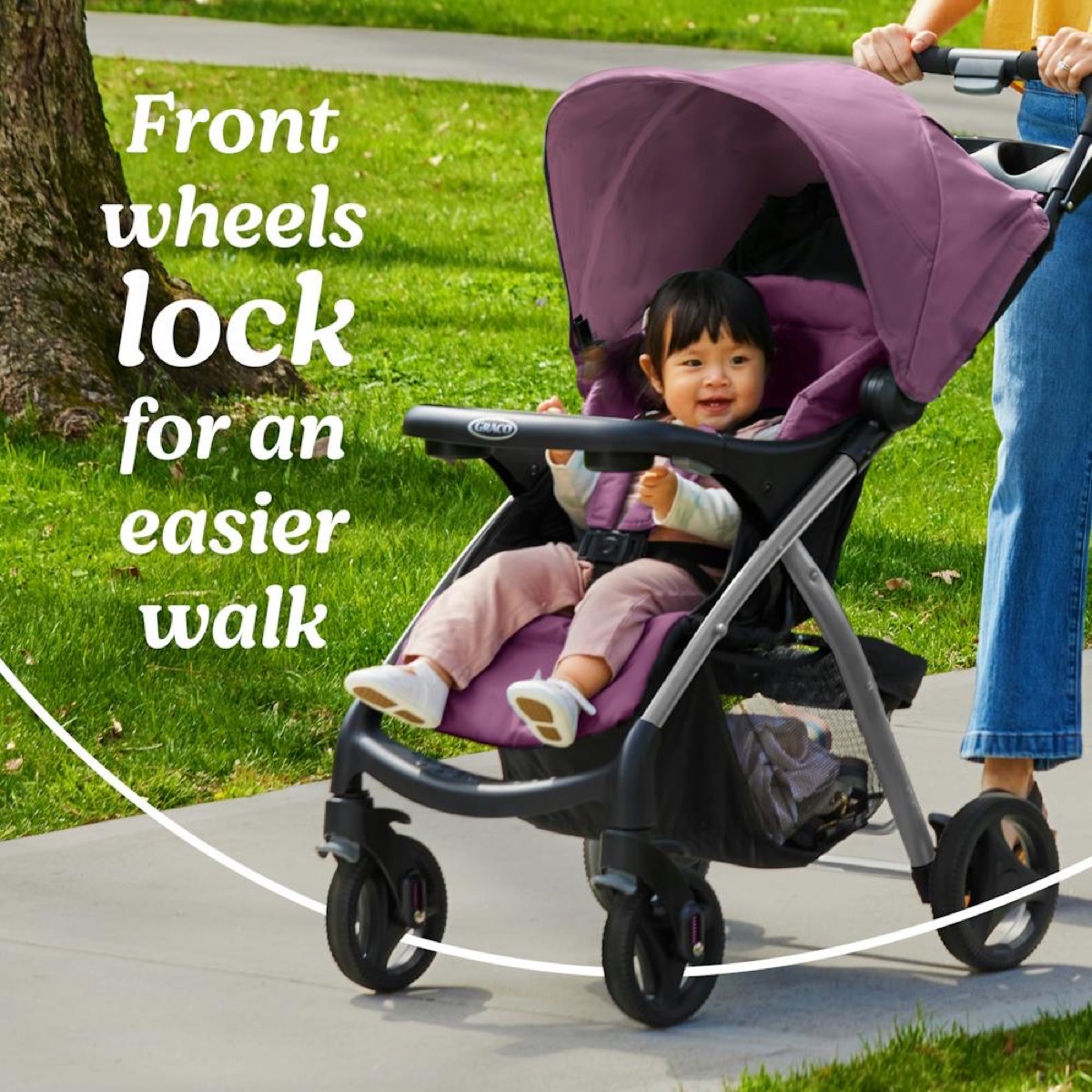 Outdoor lifestyle photo: a caregiver walks a paved park path pushing the unfolded Verb stroller with the toddler seat occupied and canopy extended, front wheel shown in locked position; overlaid text 'Front wheels lock for an easier walk'