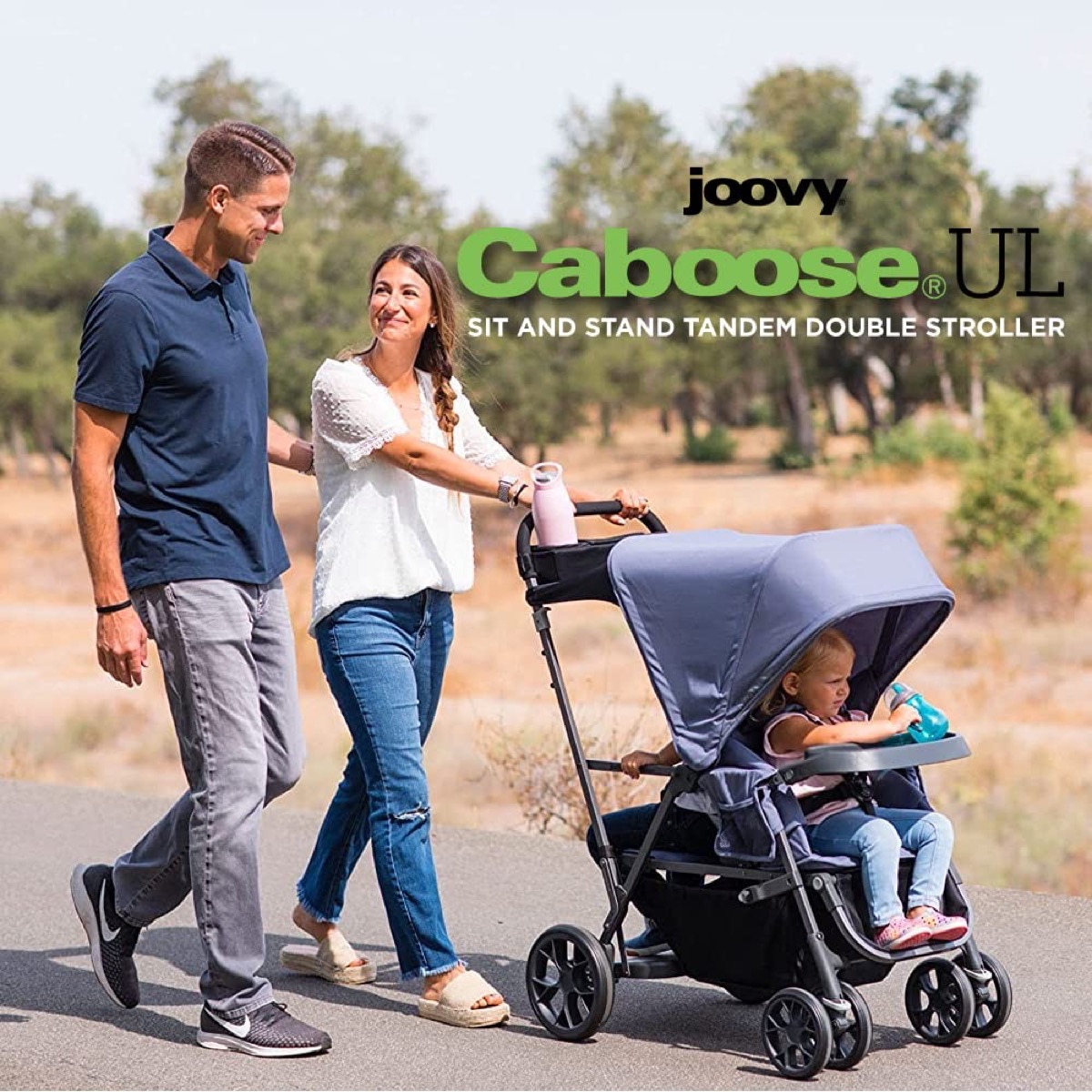 Outdoor lifestyle photo of a woman pushing the Caboose UL with a young child riding in the front seat while a man walks alongside on a paved path, with the burned-in headline 'Caboose UL — Sit and Stand Tandem Double Stroller'