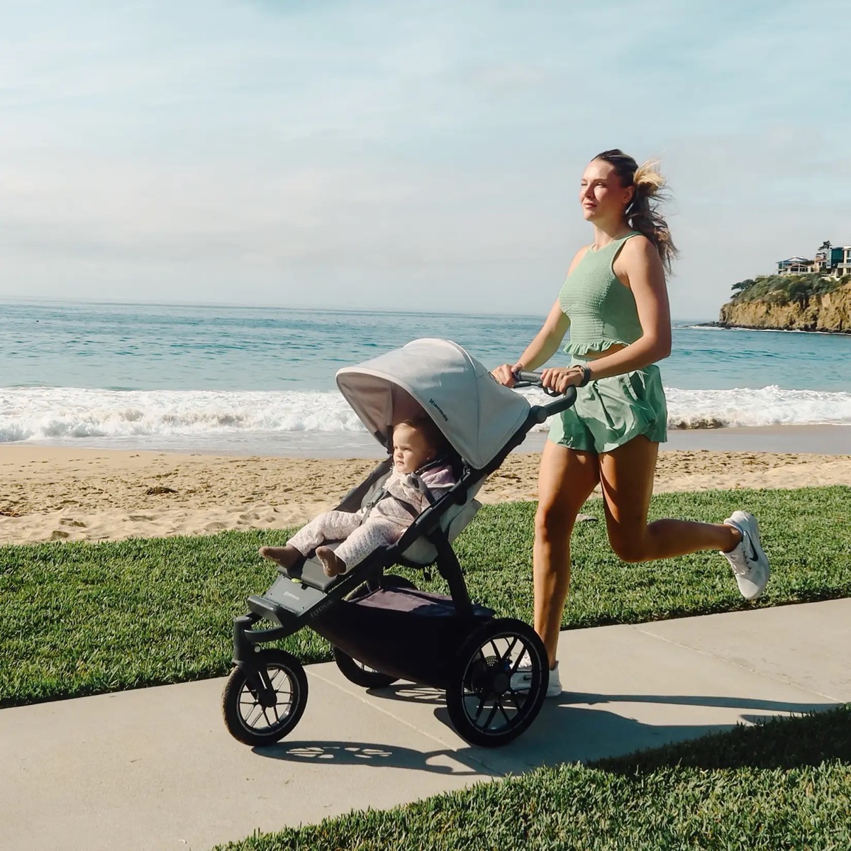A woman jogging beside the sea pushing the light-grey-canopied UPPAbaby Ridge V2 with a toddler seated in the reclined seat